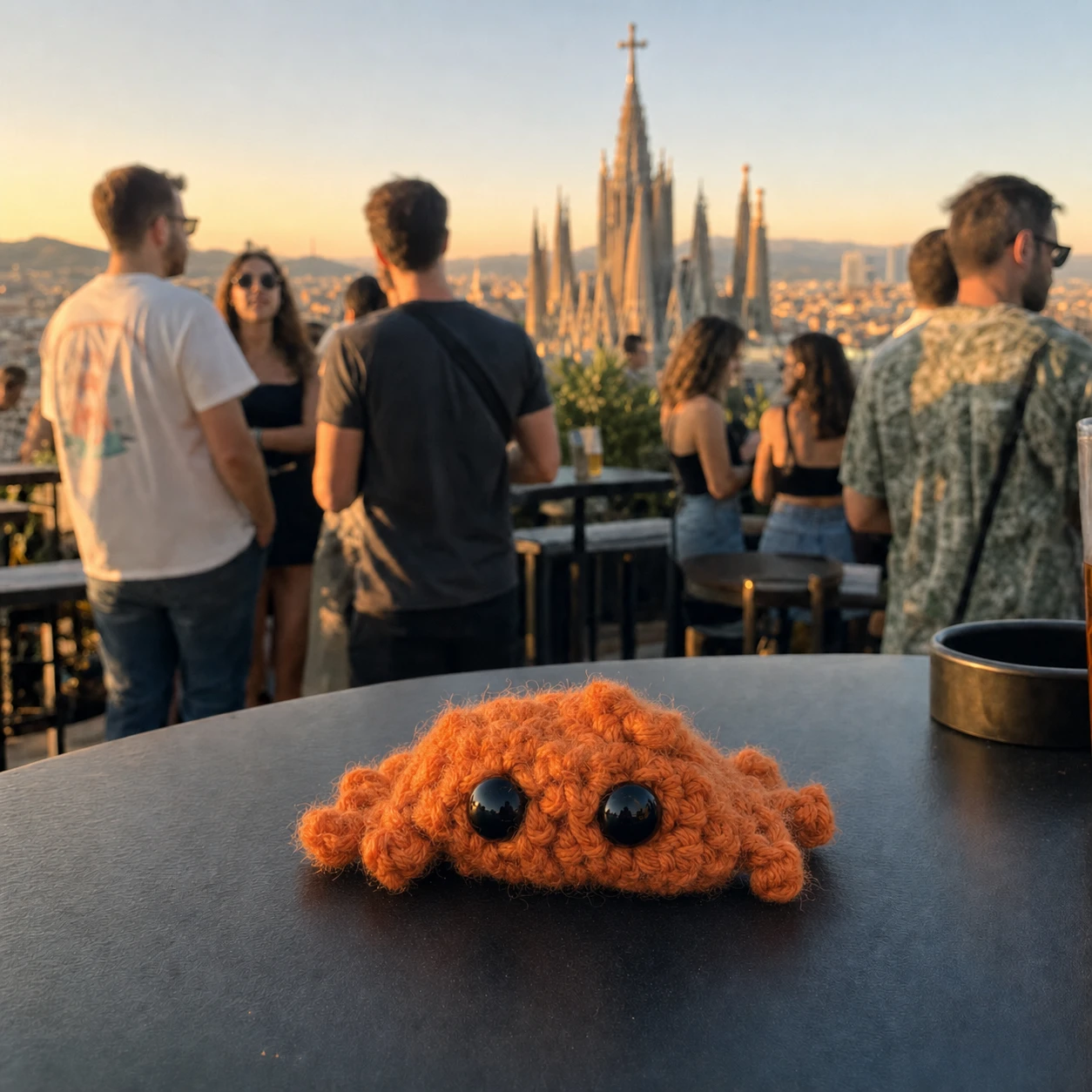 The finished Clawd, crocheted in orange with black safety eyes, sitting on a
              rooftop bar table in Barcelona with the Sagrada Família behind him at sunset.